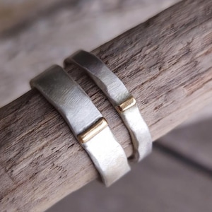 Op de afbeelding: Twee zilveren ringen met een matte, geborstelde afwerking, elk voorzien van een kleine, horizontale goudkleurige inlegbalk. De ringen hebben een licht onregelmatige, gehamerde textuur en liggen naast elkaar op een stuk drijfhout.