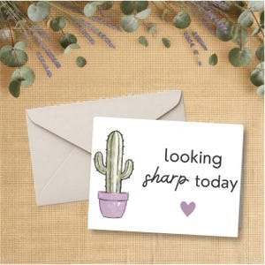 May include: A white greeting card featuring a green cactus in a light purple pot, the text "looking sharp today" in black, and a small purple heart. The card is positioned in front of a light-colored envelope on a textured tan surface.