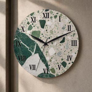 May include: A round wall clock featuring a split design with a dark green marble pattern on the left and a light-colored terrazzo pattern on the right. The clock face displays black Roman numerals and three thin black clock hands for timekeeping.