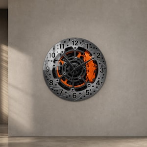 Puede incluir: Un reloj de pared circular diseñado para parecerse a un disco de freno de automóvil metálico. La esfera presenta un disco perforado de tono plateado, una estructura central negra similar a un engranaje, una pinza de freno naranja y números negros.