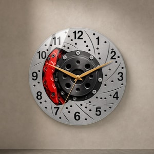Puede incluir: Un reloj de pared circular diseñado para parecer un disco de freno de automóvil plateado con una pinza de freno roja. La esfera presenta números negros del 1 al 12, un cubo central negro y tres manecillas finas de color dorado para las horas.
