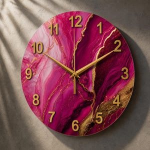 Puede incluir: Un reloj de pared redondo con un vibrante patrón de mármol magenta y rosa con vetas doradas. La esfera muestra números arábigos dorados en relieve del 1 al 12 y manecillas de hora, minuto y segundo a juego en color dorado.