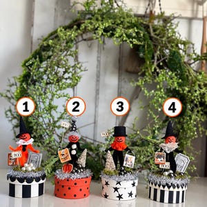 May include: Four decorative Halloween figurines featuring orange pumpkin heads on round bases. Each figure wears a hat and holds a small sign with text like BOO. The bases are decorated with patterns including black and white stripes, red dots, and black stars.