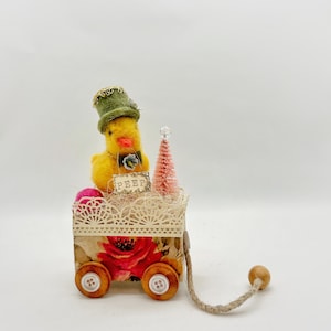 May include: A yellow felt chick wearing a green hat and holding a sign that says "PEEP" is sitting in a miniature wagon with a pink tree and pink and white yarn balls. The wagon has a floral design and wooden wheels with buttons.