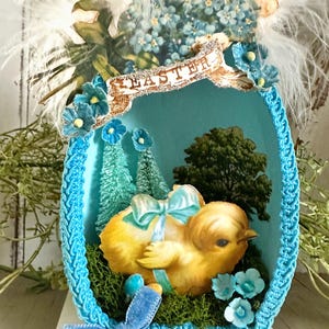 May include: An Easter-themed decorative egg with a yellow chick, blue accents, and the word "EASTER". The egg is decorated with blue flowers, a blue bow, and a textured blue border. White feathers and floral elements adorn the top.