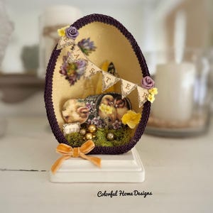 May include: A purple and gold Easter egg with a banner that reads "Easter" and a small nest with gold and white eggs inside. The egg is decorated with flowers and a bow.