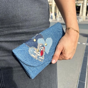 May include: A blue denim wallet featuring a flap closure adorned with two heart-shaped patches. One patch is light blue denim, while the other is dark blue denim with intricate white and red embroidery. Two small gold-toned metal studs accent the design.