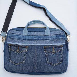 May include: A rectangular laptop bag constructed from repurposed blue denim fabric. The bag features two front pockets with buttoned flaps, a top zipper closure, two short carrying handles, and an adjustable shoulder strap made of denim.