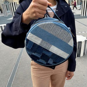 May include: A round denim bag featuring a patchwork design with various shades of blue fabric. The bag has a top handle, a silver-toned zipper closure, and a woven texture on the lower section, constructed from multiple denim panels.