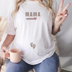 May include: A white short-sleeved t-shirt featuring the text MAMA in brown letters above a loading bar graphic with a small pink heart. Below the graphic, there are two small brown baby footprint icons centered on the lower front of the shirt.