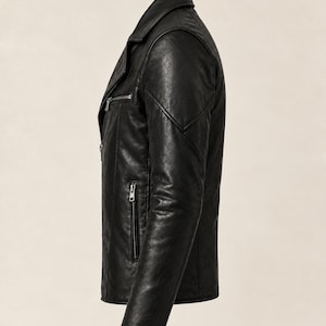 May include: A black jacket with a textured finish, featuring a notched collar, long sleeves, and silver-toned metal zippers on the front chest and side pockets. The garment is shown from a side profile view, highlighting the structured shoulder seams.