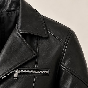 May include: A black leather jacket featuring a notched lapel collar, a horizontal zippered chest pocket with a silver-toned metal zipper pull, and visible seam detailing along the shoulder and chest area, showcasing a smooth, textured finish.