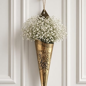 May include: A cone-shaped brass wall vase with an embossed floral design, suspended by a jute cord from a brass fleur-de-lis hook. The vase is filled with a dense arrangement of small white baby's breath flowers, creating a textured display.
