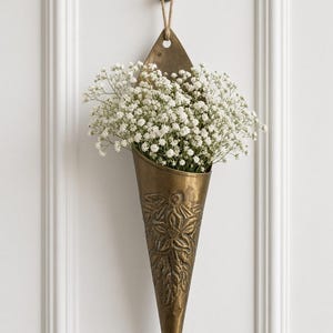 May include: A cone-shaped antique brass wall vase featuring an embossed floral design, suspended by a jute cord from a matching fleur-de-lis wall hook. The metal vessel is filled with a dense arrangement of small, delicate white baby's breath flowers.