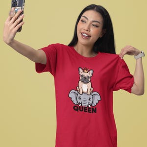 Może przedstawiać: Czerwony t-shirt z krótkim rękawem z grafiką przedstawiającą beżowego buldoga francuskiego w małej złotej koronie, siedzącego na szarym słoniu. Pod grafiką słonia znajduje się napis QUEEN wykonany pogrubionymi czarnymi wielkimi literami.