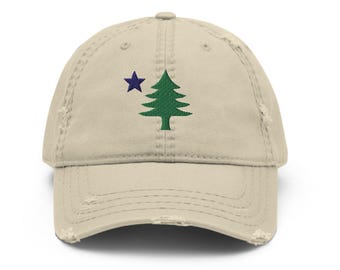 Puede incluir: Una gorra de béisbol de color beige con un acabado desgastado. El panel frontal presenta un diseño bordado que consiste en un pino verde situado a la derecha de una pequeña estrella de cinco puntas de color azul oscuro. La gorra tiene una visera curva.