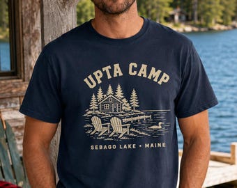 Puede incluir: Una camiseta de manga corta azul marino con un estampado gráfico color crema de una cabaña, dos sillas, un muelle y un colimbo en un lago. El texto UPTA CAMP y SEBAGO LAKE • MAINE aparece en una fuente sans-serif limpia sobre la ilustración.