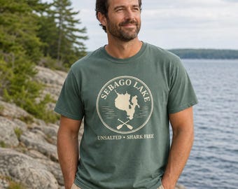 Puede incluir: Una camiseta de manga corta en color verde salvia con un gráfico circular que incluye el texto SEBAGO LAKE, un mapa, remos cruzados y el texto UNSALTED • SHARK FREE en tinta color crema. La prenda tiene cuello redondo.