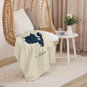 Peut inclure: Un plaid de couleur crème présentant une silhouette cartographique bleu marine foncé de l'État du Maine. Le motif comprend le mot Maine imprimé dans une police cursive bleu marine foncé, placé sous la carte avec des lignes horizontales.