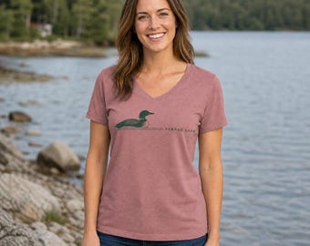Puede incluir: Una camiseta de color malva jaspeado con cuello en V que presenta un gráfico verde oscuro de un colimbo nadando en el agua, acompañado por el texto "SEBAGO LAKE" en una fuente sans-serif sencilla situada a la derecha de la ilustración.