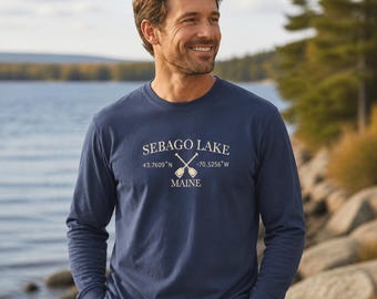 Puede incluir: Una camiseta azul marino de manga larga con cuello redondo que presenta el texto blanco SEBAGO LAKE, 43.7609°N -70.5256°W y MAINE, junto a un gráfico de dos remos cruzados situados entre las coordenadas y el nombre del estado.