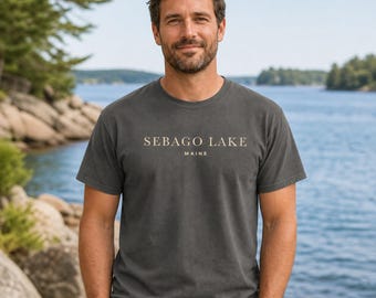 Puede incluir: Una camiseta de manga corta en color gris carbón con el texto SEBAGO LAKE MAINE impreso en una fuente serif de color beige claro en el pecho. La prenda tiene cuello redondo y un corte estándar, confeccionada en tela de algodón lisa.