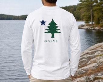 Puede incluir: Una camiseta blanca de manga larga con un gráfico en la espalda que muestra una estrella azul oscuro, un pino verde y la palabra MAINE impresa en letras mayúsculas de color azul oscuro. Tiene cuello redondo y mangas largas con puños.