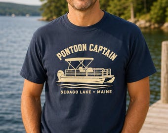 Puede incluir: Una camiseta azul marino de manga corta con un gráfico de color crema de un barco pontón. El texto PONTOON CAPTAIN está impreso sobre el barco, y SEBAGO LAKE • MAINE aparece debajo en una tipografía clara y sin remates.