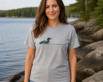 Puede incluir: Una camiseta gris jaspeado de manga corta con un gráfico de un colimbo verde oscuro nadando en el agua. El diseño incluye dos líneas horizontales y el texto SEBAGO LAKE impreso en una fuente sans-serif de color verde oscuro a la derecha.