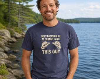 Puede incluir: Una camiseta azul marino de manga corta con el texto en color crema "WHO'D RATHER BE AT SEBAGO LAKE?" sobre un gráfico de dos manos de dibujos animados con los pulgares señalando hacia el texto "THIS GUY" centrado debajo.