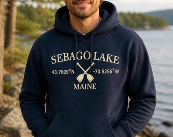 Puede incluir: Una sudadera con capucha azul marino que presenta un gráfico frontal con el texto SEBAGO LAKE, MAINE, las coordenadas 43.7609 N -70.5256 W y una ilustración de dos remos cruzados. Incluye un bolsillo tipo canguro y cordones a juego.