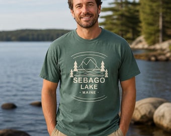 Puede incluir: Una camiseta de manga corta de color verde oscuro con un estampado gráfico en color crema que muestra un paisaje de montaña, pinos y olas de agua. El texto SEBAGO LAKE MAINE está centrado en el diseño, enmarcado por dos líneas curvas.