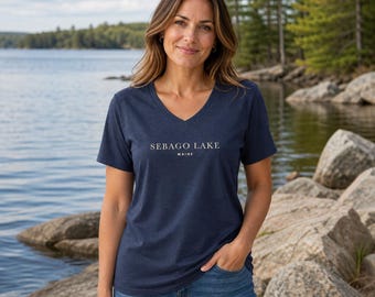 Puede incluir: Una camiseta azul marino de manga corta con cuello en V que presenta el texto blanco SEBAGO LAKE MAINE impreso en el pecho. La prenda se combina con pantalones vaqueros azules, mostrando un diseño sencillo con una tipografía centrada.