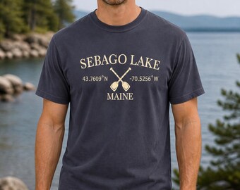 Puede incluir: Una camiseta de manga corta y cuello redondo en color gris carbón con el texto SEBAGO LAKE MAINE en crema, junto a unos remos cruzados y las coordenadas 43.7609 N y -70.5256 W impresas en el centro del pecho.