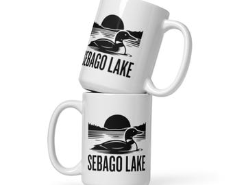 Puede incluir: Dos tazas de cerámica blanca apiladas verticalmente. Cada taza presenta un gráfico negro de un colimbo nadando en un lago con un sol poniéndose detrás de árboles. El texto SEBAGO LAKE está impreso en una fuente negra y negrita.