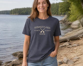 Puede incluir: Una camiseta de manga corta en color gris jaspeado oscuro con el texto SEBAGO LAKE, MAINE, las coordenadas 43.7609°N -70.5256°W y un gráfico de dos remos de madera cruzados impreso en color crema claro en el centro del pecho.