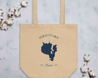 Puede incluir: Un bolso de tela beige con una silueta azul oscuro del mapa del lago Sebago, Maine. El texto SEBAGO LAKE está impreso en una fuente serif azul oscuro sobre el mapa, con la palabra Maine centrada debajo del mapa entre dos líneas horizontales.