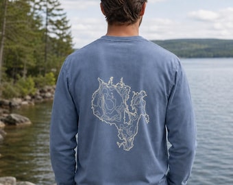 Puede incluir: Una camiseta de manga larga con cuello redondo en un color azul apagado. La parte posterior de la camiseta presenta un gráfico topográfico detallado de color claro que muestra las líneas de contorno de una masa terrestre o isla.