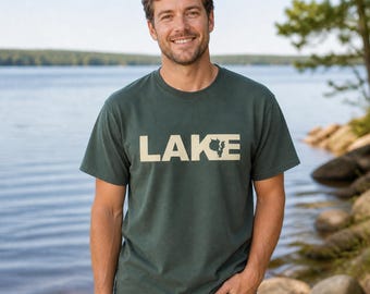 Puede incluir: Una camiseta de manga corta en color verde bosque oscuro con la palabra LAKE impresa en letras mayúsculas grandes y de color crema en el pecho, con la letra E estilizada para incorporar un pequeño contorno del estado de Wisconsin.