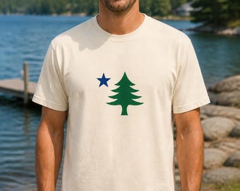 Puede incluir: Una camiseta de manga corta de color crema con un estampado gráfico en el pecho. El diseño consiste en un pino verde sólido situado a la derecha de una pequeña estrella azul de cinco puntas. La camiseta tiene un cuello redondo.