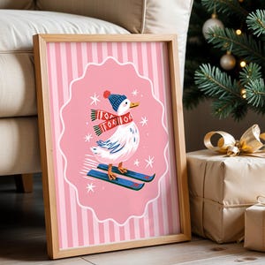 May include: A rectangular wooden frame containing a print of a white duck wearing a blue knit hat and a red and white patterned scarf while skiing on blue skis. The background of the print features pink vertical stripes and white stars.