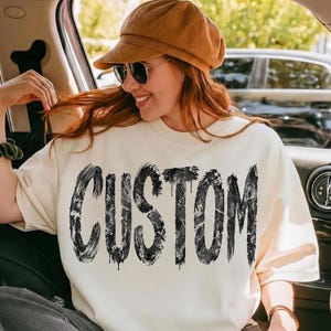 May include: A cream-coloured crewneck sweatshirt featuring the word CUSTOM printed in a distressed, black, brushstroke-style font. The garment is worn with a brown corduroy newsboy cap and dark sunglasses, showcasing a casual, relaxed fit.