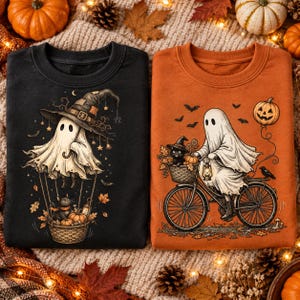 Könnte beinhalten: Ein schwarzes Sweatshirt mit einem Geist im Hexenhut im Heißluftballon mit Katze und Kürbissen, und ein oranges Sweatshirt mit einem Geist auf einem Fahrrad, einer Katze im Korb, einer Laterne und einem schwebenden Kürbisballon.
