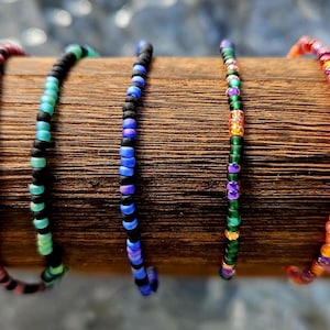 May include: A collection of seven thin beaded bracelets arranged in a row. The bracelets feature various color combinations including yellow and black, pink and black, teal and black, blue and black, green and purple, orange and red, and pink.