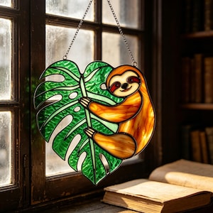 Op de afbeelding: Een glas-in-lood zonnevanger met een bruine luiaard die zich vastklampt aan een groot, levendig groen monstera-blad. Het object is geconstrueerd met donkere loden lijnen en bevat een dunne metalen ketting aan de bovenkant om op te hangen.