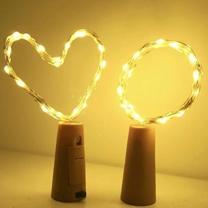 May include: Two bottle stopper lights with brown cork-shaped bases. One features a flexible wire string light shaped into a heart, and the other features a circular wire string light. Both emit a warm yellow glow from small LED bulbs.