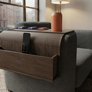 May include: A dark wood armrest tray with a ribbed top surface, a flat top section holding an orange beverage can, and a side pocket containing a black remote control. The tray is draped over the arm of a grey textured fabric armchair.