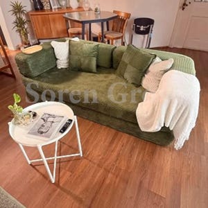 May include: An olive green corduroy sofa featuring a low back, rounded armrests, and several square throw pillows in matching green and cream colors. A white throw blanket is draped over one side. A white round side table sits in the foreground.