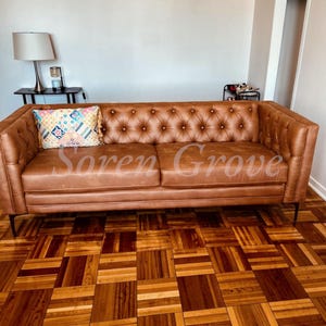 May include: A tan leather sofa featuring deep button tufting on the backrest and inner arms, supported by thin black metal legs. A rectangular throw pillow with a colorful geometric pattern rests on the left side of the seat cushions.