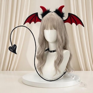 Pode incluir: Um manequim branco com uma peruca longa e ondulada loiro acinzentado, uma gargantilha preta com tachas e uma tiara com chifres de diabo vermelhos com acabamento em pelo sintético preto e asas de morcego vermelhas. Uma cauda preta longa está anexada.